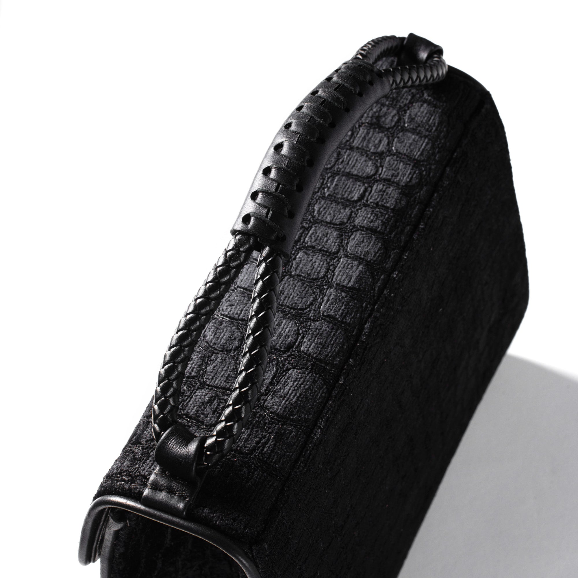 A close-up of a black textured handbag featuring a crocodile-embossed pattern, a braided leather handle, and a black metal ring clasp, set against a plain white background.
